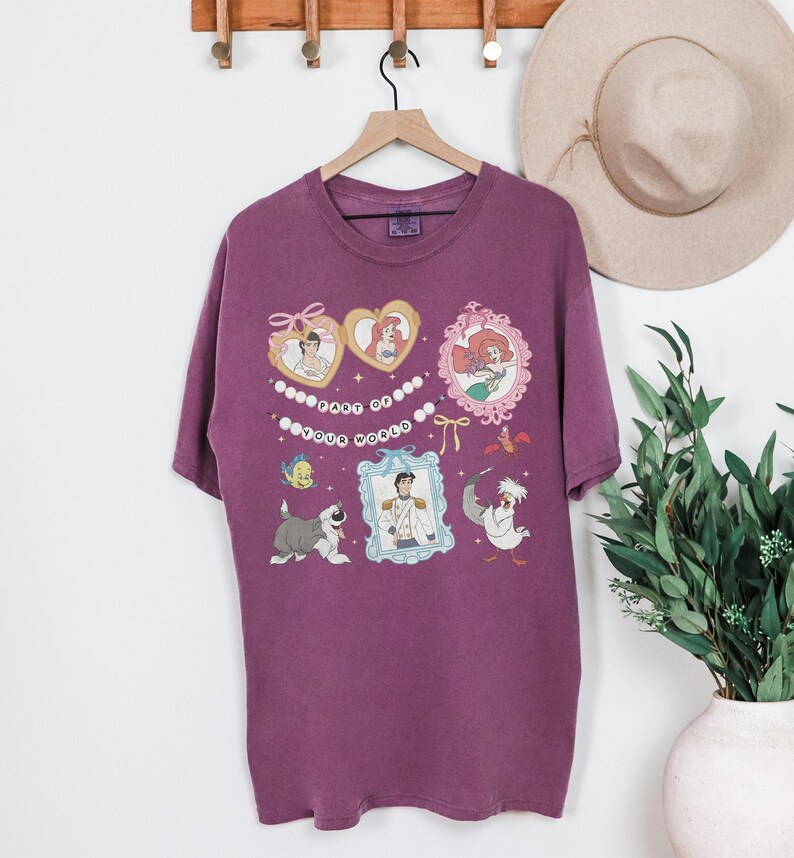 A purple t-shirt with a Disney-themed design hanging on a wooden hanger, along with a beige hat and a potted plant.