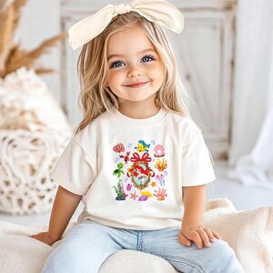 A young girl with blonde hair wearing a white t-shirt with a colorful underwater-themed design, smiling at the camera.