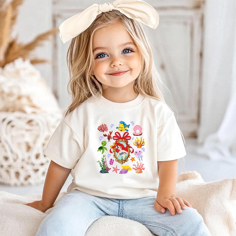 A young girl with blonde hair wearing a white t-shirt with a colorful underwater-themed design, smiling at the camera.