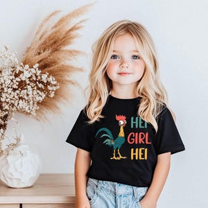 A young girl with blonde hair is wearing a black t-shirt with a colorful graphic of a rooster and the text "HEE GIRL HEI" printed on it. She is standing in front of a vase with white flowers, and there is a wooden table with a white vase on it.