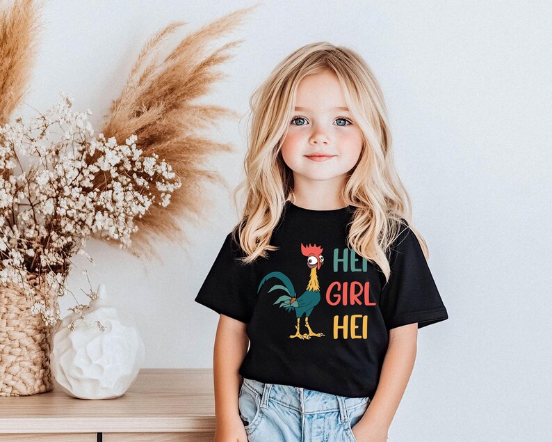 A young girl with blonde hair is wearing a black t-shirt with a colorful graphic of a rooster and the text "HEE GIRL HEI" printed on it. She is standing in front of a vase with white flowers, and there is a wooden table with a white vase on it.