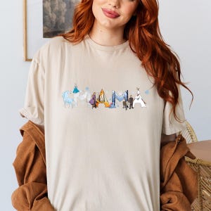 A woman with long red hair wearing a beige t-shirt with a graphic design of various characters from the Disney movie Frozen.