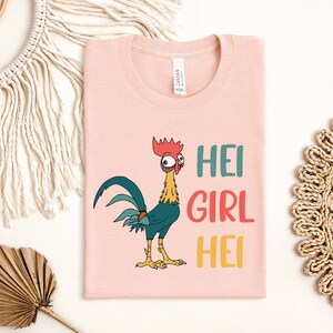 a pink t-shirt with a cartoon rooster and the text "HEI GIRL HEI" printed on it. The t-shirt is laid out on a white surface, surrounded by various decorative items such as a woven mat, a leaf, and a string.