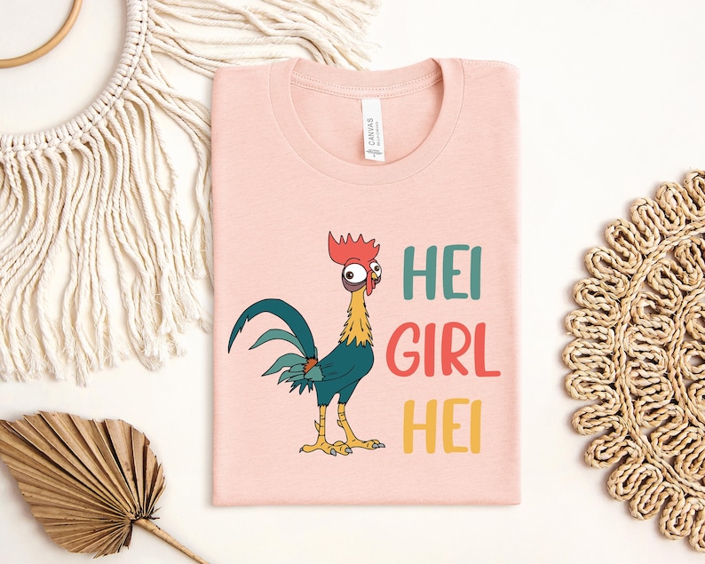 a pink t-shirt with a cartoon rooster and the text "HEI GIRL HEI" printed on it. The t-shirt is laid out on a white surface, surrounded by various decorative items such as a woven mat, a leaf, and a string.