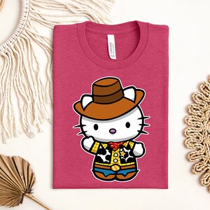 A pink t-shirt with a cartoon image of a cat wearing a cowboy hat and a yellow and blue outfit, along with a straw hat and a woven mat.