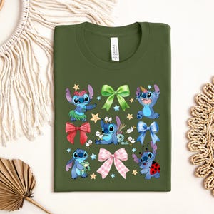 a green t-shirt with a colorful design of cartoon characters, including Stitch, a blue bunny, and other whimsical elements. The t-shirt is displayed on a white background, surrounded by various decorative items such as a woven basket, a leaf, and a star.