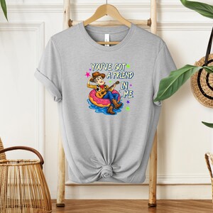 A gray t-shirt with a cartoon character playing a guitar and the text "You've Got a Friend in Me" printed on it, hanging on a wooden hanger.