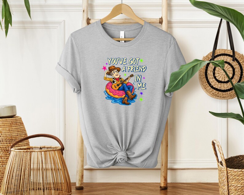 A gray t-shirt with a cartoon character playing a guitar and the text "You've Got a Friend in Me" printed on it, hanging on a wooden hanger.