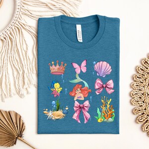 a blue t-shirt with a colorful graphic design depicting various sea-themed elements, including a mermaid, crown, butterfly, shell, bow, and coral.