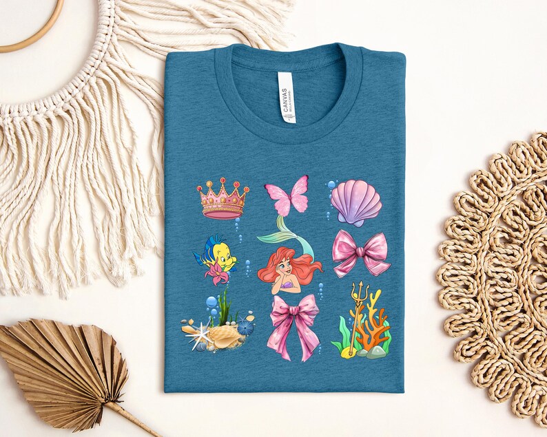 a blue t-shirt with a colorful graphic design depicting various sea-themed elements, including a mermaid, crown, butterfly, shell, bow, and coral.