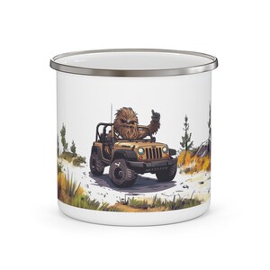 May include: White enamel mug with a brown and white illustration of a furry creature driving a jeep on a dirt road. The creature is wearing a brown shirt and has a brown beard. The jeep is brown and has a white roof.