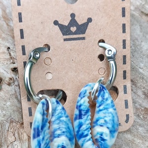 May include: A pair of silver hoop earrings with blue and white seashell charms. The earrings are displayed on a brown card with a crown logo.