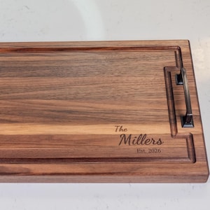 May include: A rectangular dark brown wooden serving tray with a natural wood grain pattern. The tray features two black metal handles on the sides and a juice groove. Engraved on the surface is the text "The Millers Est. 2026" in a script font.