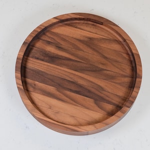 May include: A round wooden serving board with a raised edge. The board is made of dark brown wood with visible grain patterns. The board is approximately 12 inches in diameter and sits on a white surface.