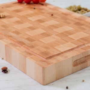 May include: A thick, rectangular wooden cutting board with a light tan, end-grain checkerboard pattern. The board features a recessed juice groove along the perimeter and integrated side handles carved directly into the wood block.
