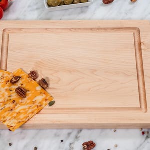 May include: A rectangular light-colored wooden cutting board with a smooth surface and a carved juice groove along the perimeter. The board is positioned horizontally and features a natural wood grain pattern throughout the flat surface.