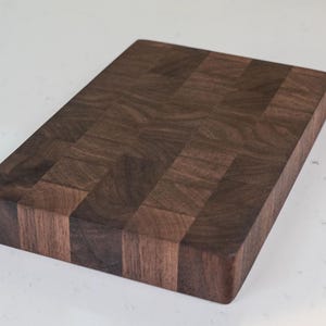 Puede incluir: Tabla de cortar rectangular de madera marr&oacute;n oscuro. El tablero tiene un patr&oacute;n de bloques y est&aacute; sobre un fondo blanco. La veta de la madera es visible y los bordes son lisos.