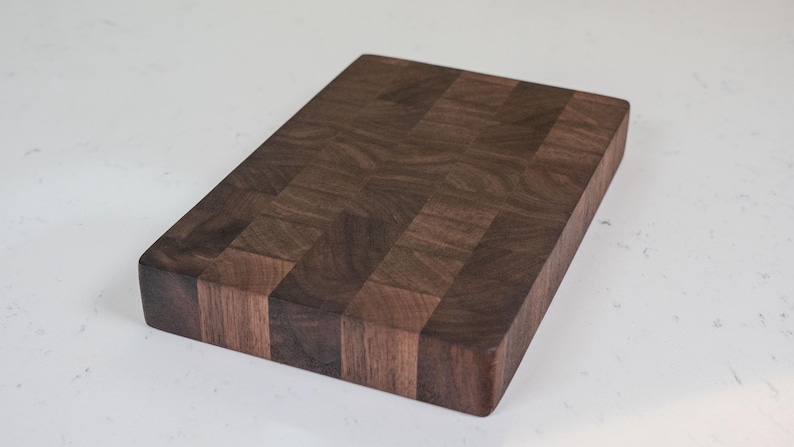 Puede incluir: Tabla de cortar rectangular de madera marr&oacute;n oscuro. El tablero tiene un patr&oacute;n de bloques y est&aacute; sobre un fondo blanco. La veta de la madera es visible y los bordes son lisos.