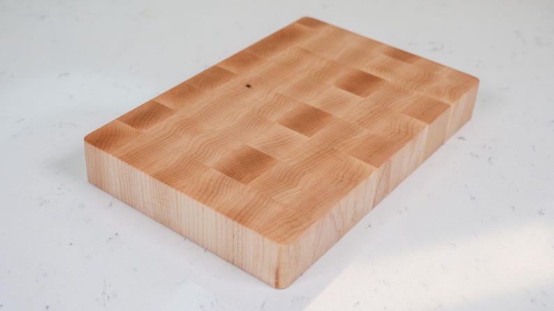 Puede incluir: Una tabla de cortar rectangular de grano final. La tabla est&aacute; hecha de madera de color claro con un patr&oacute;n de tablero de ajedrez. La veta de la madera es visible y la tabla tiene bordes redondeados. Este art&iacute;culo de cocina es ideal para la preparaci&oacute;n de alimentos.