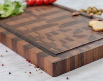 May include: A rectangular end-grain wooden cutting board featuring a dark and medium brown checkered pattern. The board has a deep juice groove carved along the perimeter and thick, sturdy edges. It is displayed with scattered peppercorns.