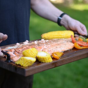 May include: A wooden serving board holds a grilled salmon fillet, corn on the cob, and colorful bell peppers. The salmon is seasoned and cooked to perfection. The board is being held by a person.