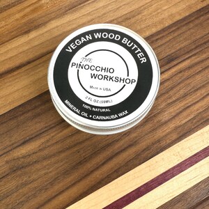 May include: A round, silver tin of vegan wood butter labeled "The Pinocchio Workshop Vegan Wood Butter" with the text "Made in USA, 2 fl oz (59 ml), 100% Natural, Mineral Oil + Carnauba Wax".