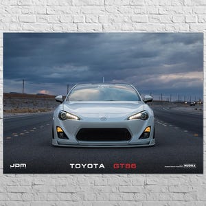以下が含まれることがあります： 白いトヨタGT86スポーツカーが道路の中央に配置され、ヘッドライトが点灯しています。画像には「JDM」と「TOYOTA GT86」の文字が赤と白で表示されています。背景には曇り空と砂漠の風景が写っています。
