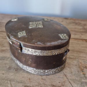 May include: An oval-shaped, antique brass trinket box with a silver-coloured trim and decorative accents. The box has a hinged lid and a small latch.