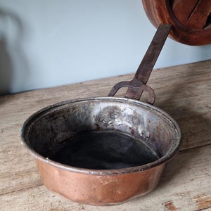 May include: An antique copper pan with a long, riveted iron handle. The pan has a rounded bowl shape with a dark interior and a tarnished exterior. A wooden object is partially visible in the background.
