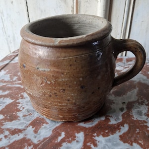 Può includere: Una tazza in ceramica marrone con manico. La tazza ha una forma leggermente irregolare e una superficie strutturata. L'interno è di un marrone più scuro. È appoggiata su una superficie con motivi rossi e bianchi.