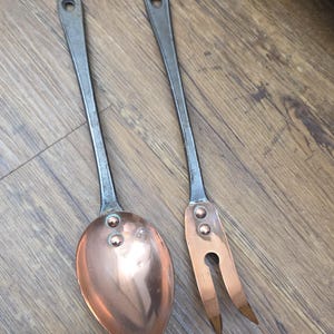 May include: A copper spoon and fork set with riveted construction. The spoon has a rounded bowl, and the fork has two tines. Both utensils have long, slender handles with a hole at the end for hanging. The metal has a rustic, aged appearance.