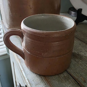 May include: A brown ceramic mug with a smooth, cylindrical body and a single curved handle. The exterior features a matte, earthy brown finish with subtle horizontal ridges, while the interior is a contrasting light, off-white glazed color.