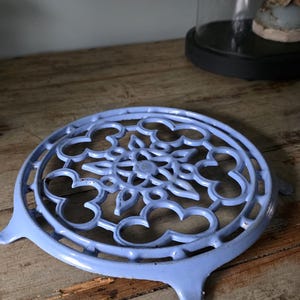 May include: A light blue, cast iron trivet with a floral design. The trivet has a circular shape with a decorative pattern and three small feet. It is placed on a rustic wooden surface.