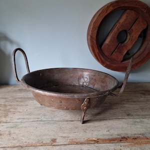 May include: A vintage copper pan with two curved handles and three small feet. The pan is round and has a dark, aged appearance. A wooden wheel-like object is in the background, giving a rustic feel to the image.