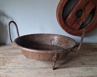 May include: Antique copper pan with two arched handles and three small legs. The pan has a round shape and a dark, aged patina. A wooden wheel-like object hangs in the background, adding a rustic touch to the scene.