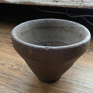 May include: A dark brown, conical-shaped bowl with a rough, textured surface and a wide, open rim. The exterior and interior of the bowl feature a weathered, matte finish with visible imperfections and a small chip along the top edge of the rim.