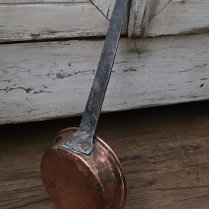 May include: A round copper pan featuring a long, dark, textured metal handle attached with two visible rivets. The pan has a polished, warm metallic finish and a flat base, with a small hole at the top of the handle for hanging purposes.