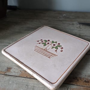 May include: Square ceramic trivet with a floral design. The trivet features a cream-colored surface with a brown border and a central illustration of flowers in a basket, with green leaves and red berries.