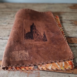 Puede incluir: Una funda de cuero marrón con un borde cosido. La funda presenta una imagen en relieve de una catedral. El forro interior tiene diseños florales en naranja, amarillo y blanco. La funda está doblada y apoyada sobre una superficie de madera.