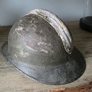 May include: An antique, weathered metal helmet with a curved, reinforced brow. The helmet has a dark, matte finish with visible signs of wear and age, resting on a rustic wooden surface. The helmet's design suggests a historical or military context.