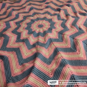 Könnte beinhalten: Handgefertigte Häkeldecke mit Sternmuster. Die Decke hat abwechselnde Streifen in Hellblau, Rosa und Creme. Die Decke ist weich und gemütlich, perfekt zum Einkuscheln auf der Couch oder im Bett.