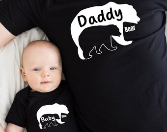 May include: A black t-shirt with the text "Daddy Bear" and a white bear silhouette. A matching black bodysuit with the text "Baby Bear" and a white bear silhouette. The shirts are worn by a father and baby.
