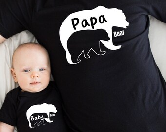 May include: A black t-shirt with a white bear outline that says "Papa Bear" and a smaller bear inside the outline. A baby is wearing a black onesie with a white bear outline that says "Baby Bear".