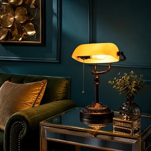 May include: A banker lamp with a curved amber glass shade and a dark metal base featuring ornate detailing. The lamp includes a pull chain switch and emits a warm glow, standing on a reflective surface next to a small decorative box.
