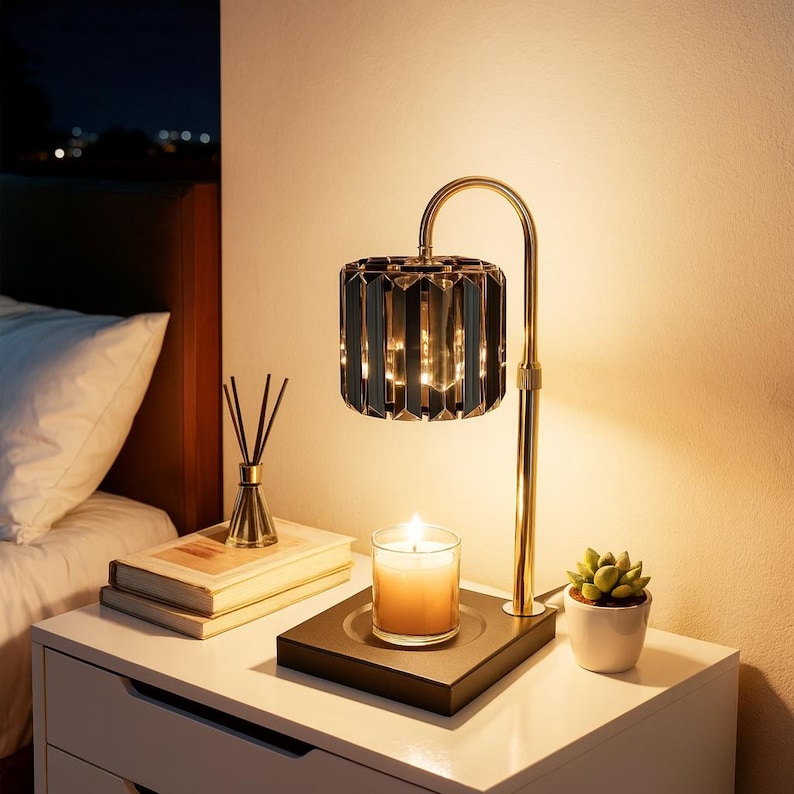 May include: A bedside table with a lit candle, a small succulent in a white pot, and a decorative lamp with a gold base and a black, faceted shade. A stack of books and a reed diffuser are also on the table.