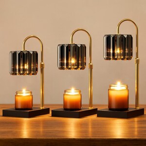 May include: Three candle holders with a modern design. Each has a black base, a gold-colored arched support, and a faceted, dark gray glass shade. A lit candle sits beneath each shade, casting a warm glow. The holders are arranged on a wooden surface.