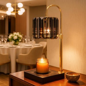 May include: A decorative table lamp with a faceted, smoky-gray glass shade and a gold-colored metal frame. A lit candle in a glass jar sits on a black base below the lamp. A small, textured bowl is also on the table.