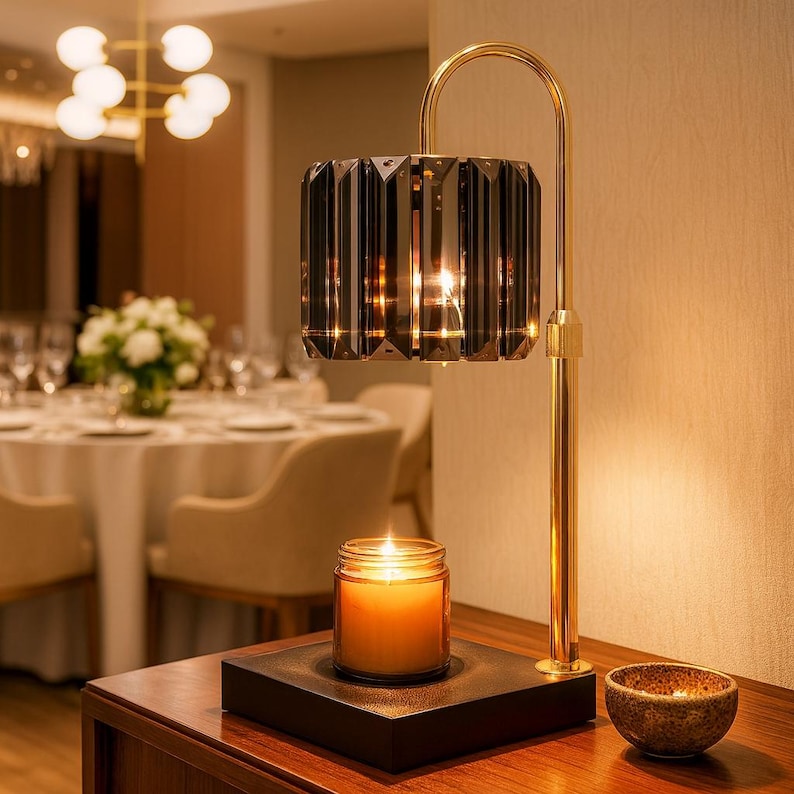 May include: A decorative table lamp with a faceted, smoky-gray glass shade and a gold-colored metal frame. A lit candle in a glass jar sits on a black base below the lamp. A small, textured bowl is also on the table.