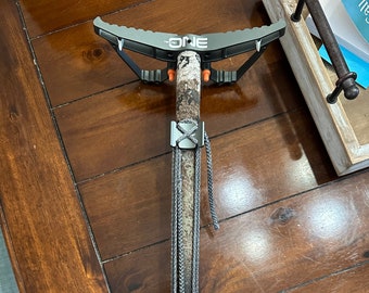 May include: A camouflage-patterned tree stand with a black metal frame and a gray rope. The stand has a metal footrest and a metal platform. The stand is labeled "The ONE" and has a black and orange safety strap.