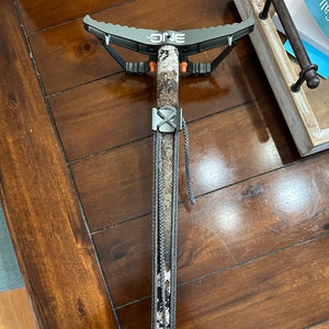 May include: A camouflage-patterned tree stand with a black metal frame and a gray rope. The stand has a metal footrest and a metal platform. The stand is labeled "The ONE" and has a black and orange safety strap.